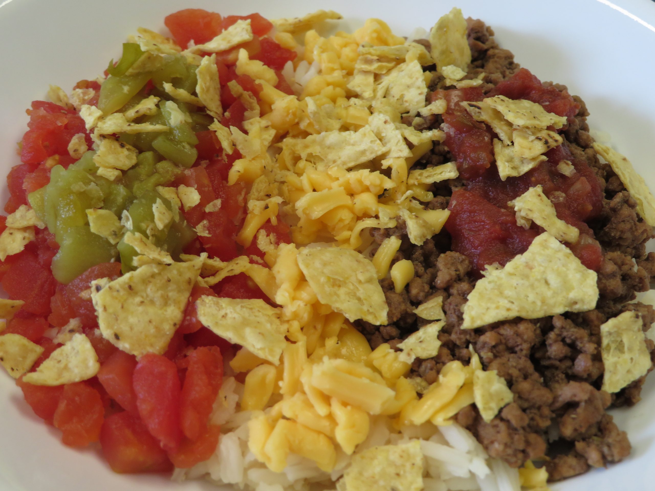 A bowl of white rice topped with hamburger, tomatoes, salsa, green chiles, cheese, and tortilla chips