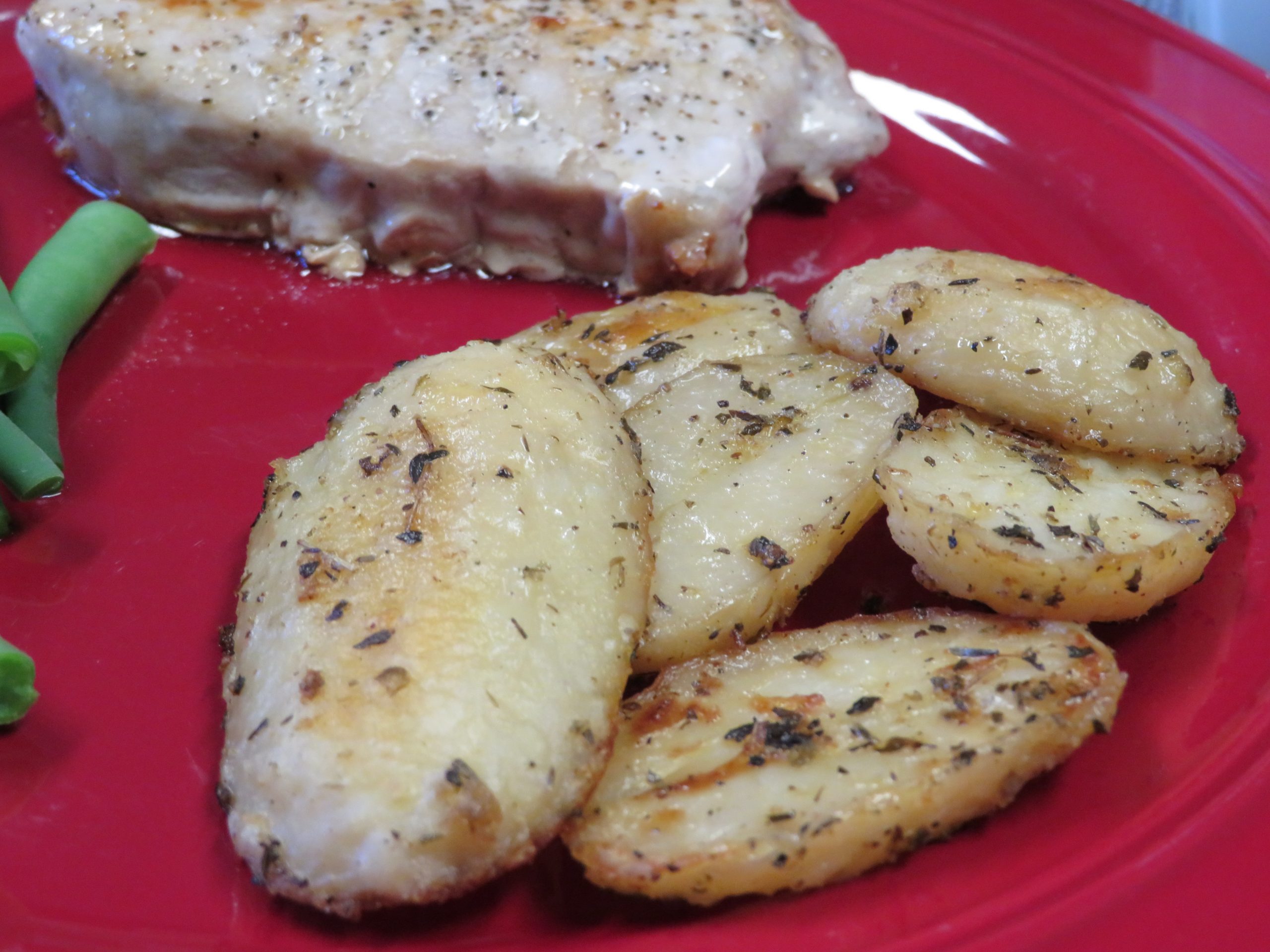 Herb roasted potatoes on a red plate with green beans and a pork chop