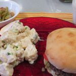 Hamburger on a red plate with potato salad and 3-bean salad in a bowl