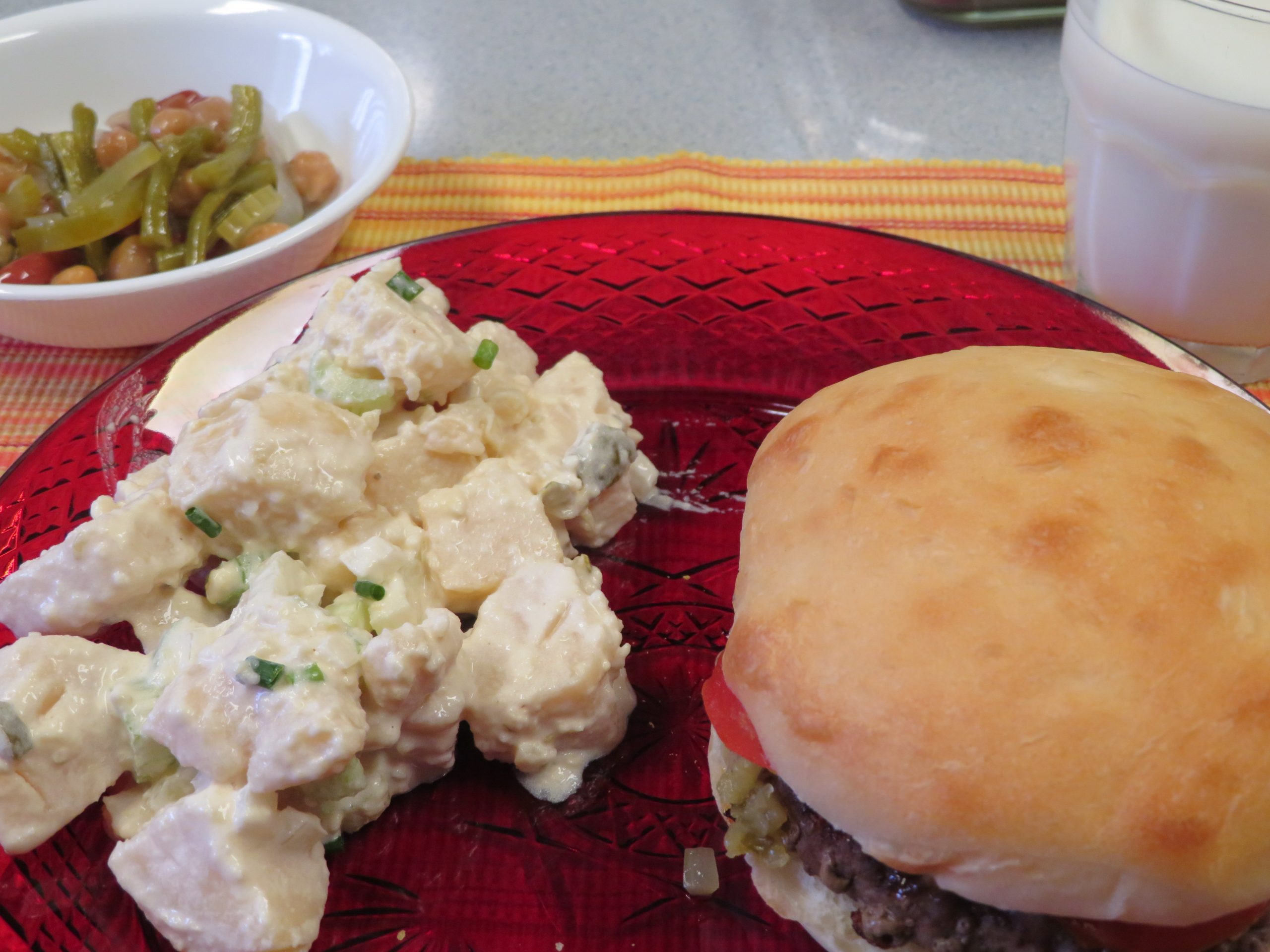 Hamburger on a red plate with potato salad and 3-bean salad in a bowl