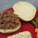 Smoked Brisket sandwich on a red plate with Mashed Potato Salad, pickles, and cut up vegetables