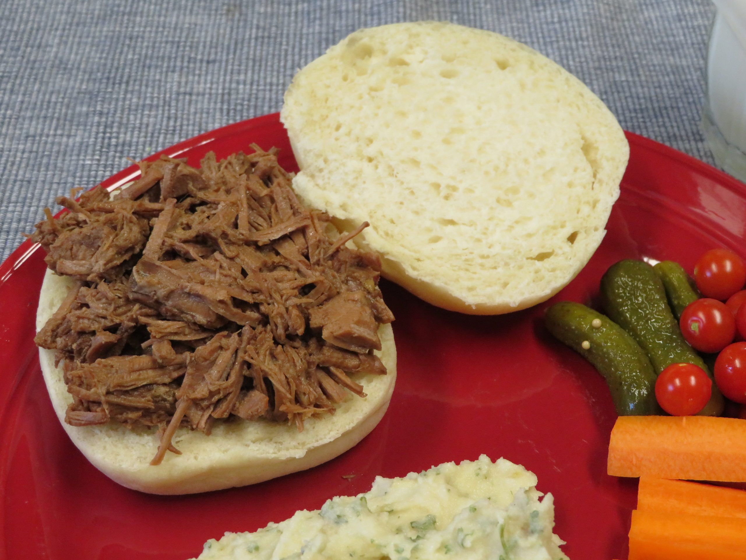 Smoked Brisket sandwich on a red plate with Mashed Potato Salad, pickles, and cut up vegetables